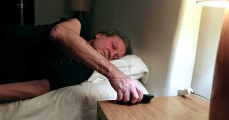 Téléchargez les photos : Man turns nightstand light ON and picks up cellphone. Person checking smartphone screen - en image libre de droit