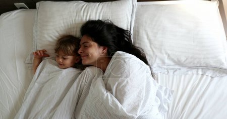 Foto de Toddler and mom playing hide and seek under blanket in bed. Peepaboo play. Mother and baby cuddling in bed together - Imagen libre de derechos