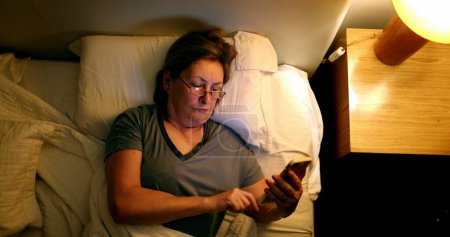 Téléchargez les photos : Older woman using smartphone device in bed looking at phone screen before sleep - en image libre de droit