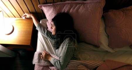 Téléchargez les photos : Single woman lying in bed turning off bedside lamp nightstand - en image libre de droit