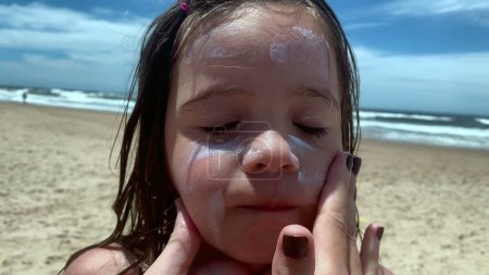 Téléchargez les photos : Parent appliquant de la crème solaire sur le visage de sa fille. Mère frottant et répandant la crème solaire à l'enfant - en image libre de droit