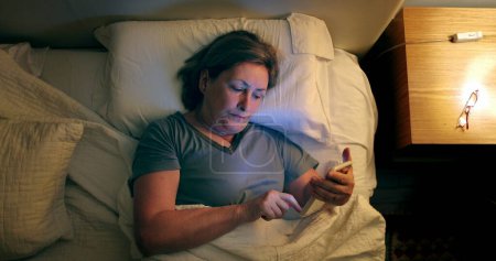 Photo for Woman turns on nightstand light and picks up cellphone in the middle of the night - Royalty Free Image