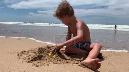 Téléchargez les photos : Petit garçon jouant au château de construction de plage avec du sable au bord de la mer - en image libre de droit