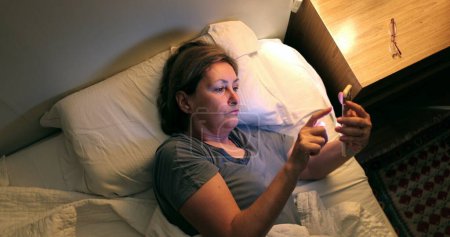 Téléchargez les photos : Woman picks up phone from nightstand lying in bed. Older lady browsing internet on smartphone before sleep - en image libre de droit