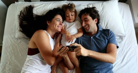 Photo for Millennial couple in bed with baby toddler infant boy watching content on cellphone device - Royalty Free Image