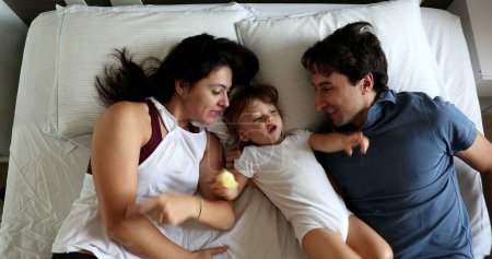 Téléchargez les photos : Happy mother and father with baby infant son in bed, top view. Toddler kissing parents in cheek, loving family caring relationship - en image libre de droit