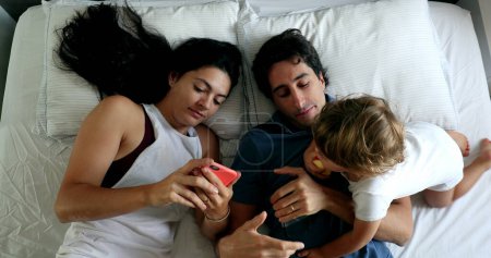 Photo for Millennial couple in bed with infant toddler boy wanting attention. Man and woman lying down looking at cellphone, top view - Royalty Free Image