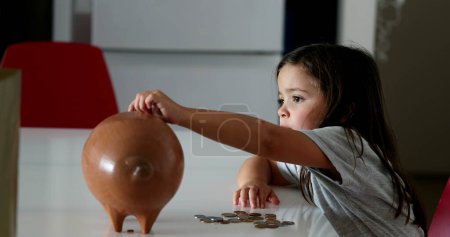 Téléchargez les photos : Little girl adding coins inside piggy bank. Child saving money concept - en image libre de droit