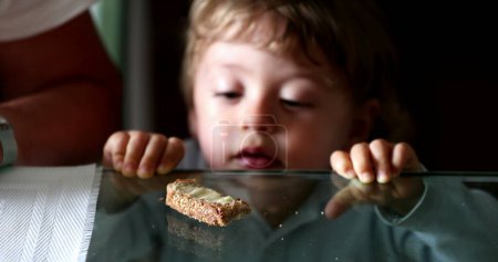 Téléchargez les photos : Child snacking bread and butter. Baby boy leaning on glass table grabbing food - en image libre de droit
