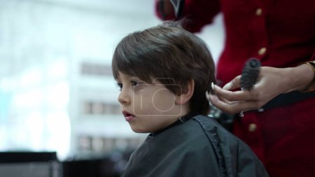 Téléchargez les photos : Petit garçon se faire couper les cheveux et peigner par un coiffeur professionnel au salon de coiffure, personne utilisant un sèche-cheveux - en image libre de droit
