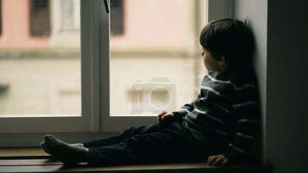 Téléchargez les photos : Petit garçon mélancolique assis par la fenêtre regardant bruine de l'appartement, enfant portant un pull regardant la vue avec une émotion contemplative coûteuse - en image libre de droit