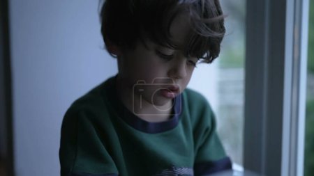 Téléchargez les photos : Triste enfant déprimé avec une expression mélancolique regardant vers le bas avec une expression sombre, assis par la fenêtre à la maison la nuit - en image libre de droit