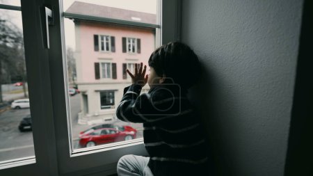 Foto de Triste niño deprimido mirando a la calle desde el segundo piso, queriendo salir, niño aburrido sin nada que hacer, apoyado en la ventana en casa - Imagen libre de derechos