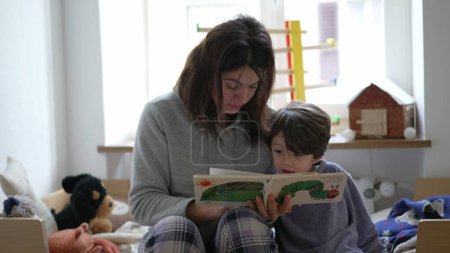 Foto de Auténtica escena de dormitorio de la madre leyendo la historia a su hijo pequeño, momento de crianza cándida con mamá y libro - Imagen libre de derechos