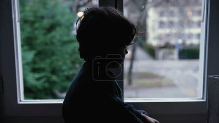 Téléchargez les photos : Enfant mélancolique assis par la fenêtre à la maison se sentant triste et déprimé - en image libre de droit