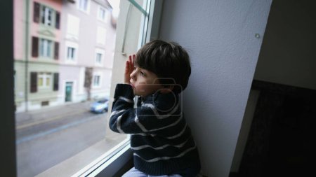 Téléchargez les photos : Un enfant ennuyé penché sur la fenêtre regardant par la fenêtre, petit garçon voulant sortir. Triste enfant déprimé sans rien faire, regardant la rue depuis la maison du deuxième étage - en image libre de droit