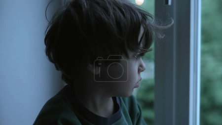 Téléchargez les photos : Enfant déprimé mélancolique assis par la fenêtre dans l'immeuble, montrant la dépression et la tristesse dans le désespoir calme - en image libre de droit