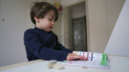 Foto de Niño pequeño leyendo libro sentado en el escritorio en el dormitorio. Niño curioso estudiando por sí mismo pasando página y mirando el texto y las imágenes - Imagen libre de derechos