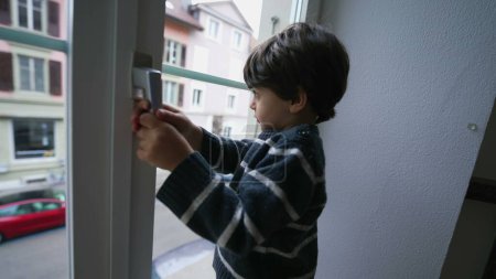 Téléchargez les photos : Un petit garçon tenant dans la poignée de bouton de fenêtre de l'appartement au deuxième étage, concept de sécurité des enfants - en image libre de droit