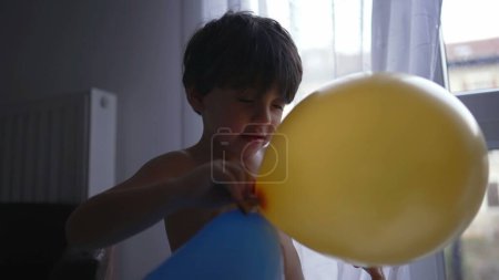 Photo for Kid blows up balloons at home after birthday party. Little boy bites balloon with teeth - Royalty Free Image
