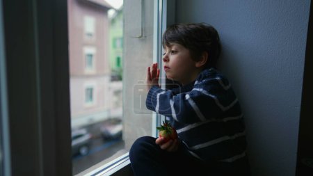 Téléchargez les photos : Mélancolique petit garçon regardant Vue de la fenêtre, voulant sortir mais coincé à la maison dans triste émotion ennuyeuse - en image libre de droit