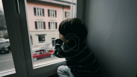 Téléchargez les photos : Triste petit garçon déprimé qui regarde la rue du deuxième étage, qui veut sortir, qui s'ennuie, qui s'appuie sur la fenêtre à la maison - en image libre de droit