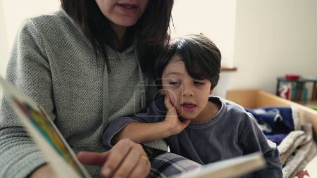 Téléchargez les photos : Une mère fiancée raconte des histoires à un enfant absorbé au chevet du lit, scène de style de vie d'un parent lisant un livre à un petit garçon - en image libre de droit