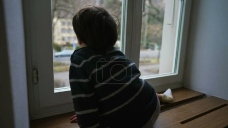 Foto de Pequeño niño pensativo mirando desde la ventana del apartamento, profundamente en el pensamiento - Imagen libre de derechos