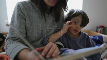 Téléchargez les photos : Mère lisant un livre à un petit garçon assis au chevet d'un lit, Enfant absorbé par l'histoire, Parent engagé dans la scène de style de vie de la maternité - en image libre de droit