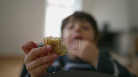 Foto de Mordedura de pan saboreador infantil de primer plano de la hora de la merienda de los jóvenes en la mesa en interiores. Niño comiendo pan - Imagen libre de derechos