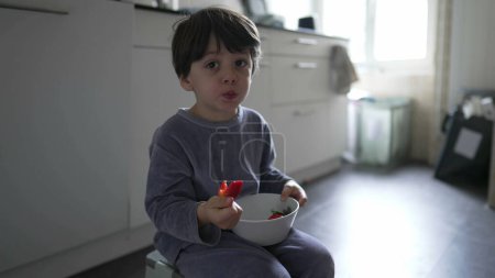 Photo for Child eating strawberries sitting on kitchen floor wearing pajamas. Little boy eats morning breakfast holding bowl in hand and tasting fruit - Royalty Free Image