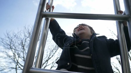 Foto de Alegre niño escalando patio corredera con chaqueta. enérgico niño pequeño haciendo ejercicio en el parque al aire libre, expresión feliz de niño subiendo - Imagen libre de derechos