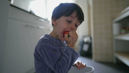 Téléchargez les photos : Petit garçon en pyjama mangeant des fraises pour le petit déjeuner, assis sur le plancher de la cuisine avec bol à la main - en image libre de droit