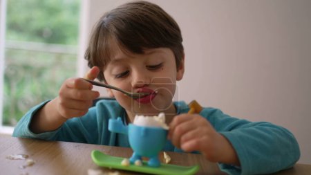 Photo for Concentrated child eating soft boiled egg in the evening, dining healthy meal with spoon by himself, wearing pajamas - Royalty Free Image
