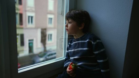 Téléchargez les photos : Ennuyé triste émotion de petit garçon assis par la fenêtre regardant la vue dans la mélancolie, enfant voulant sortir coincé à la maison - en image libre de droit