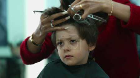 Foto de Primer plano niño conseguir un corte de pelo en la peluquería. Peluquero profesional que corta y peina el pelo del niño, expresión pensativa severa - Imagen libre de derechos