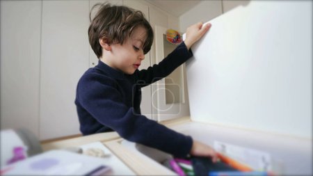 Foto de Un niño pequeño sentado en el escritorio abriendo el cajón de la mesa y tomando material de dibujo, comprometido con el trabajo de la escuela de kindergarten en casa - Imagen libre de derechos