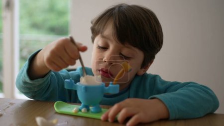 Foto de Niño comiendo huevo blando hervido para el desayuno con cuchara. Niño de 4 años comiendo solo, cara de cerca absorbida en bocadillos saludables, oeuf a la coque - Imagen libre de derechos