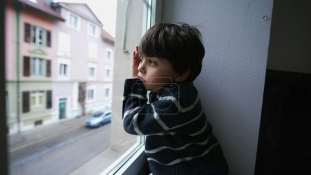 Foto de Un niño aburrido apoyado en una ventana mirando hacia afuera, un niño pequeño queriendo salir. Triste chico deprimido sin nada que hacer, mirando a la calle desde la casa del segundo piso - Imagen libre de derechos