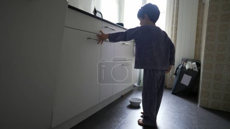 Photo for Little boy throwing away rest of food into the garbage standing inside kitchen wearing pajamas. Authentic domestic lifestyle scene of kid tidying up home - Royalty Free Image