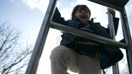 Téléchargez les photos : Joyeux enfant grimpant glissière terrain de jeu veste. petit garçon énergique faisant de l'exercice dans un parc extérieur, expression heureuse d'un enfant qui monte - en image libre de droit