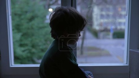 Téléchargez les photos : Triste enfant déprimé avec une expression mélancolique regardant vers le bas, assis par la fenêtre à la maison la nuit - en image libre de droit