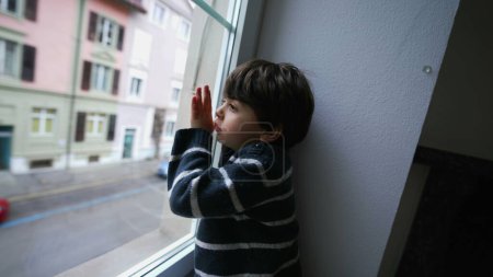 One bored child leaning on window looking out, little boy wanting to go outside. Sad depressed kid with nothing to do, staring at street from second floor home