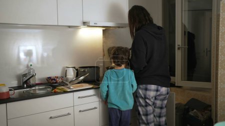 Photo for Back of mother and child cooking at kitchen, casual domestic family lifestyle scene of small boy observing mom preparing meal at night, authentic and real life - Royalty Free Image
