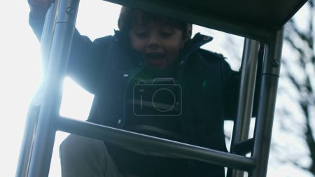 Téléchargez les photos : Enfant excité monte escaliers coulissants à l'extérieur avec rétro-éclairage sunflare. Joyeux petit garçon serrant sur des barres métalliques montant vers le haut pendant la saison d'automne - en image libre de droit