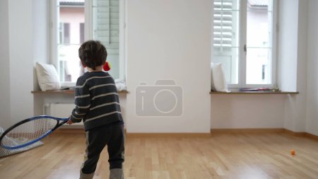 Foto de Rising Tennis Star / Child 's Tennis Juega en un apartamento vacío, la familia se traslada a un nuevo hogar. ejercicios de niño pequeño golpeando la pelota con raqueta en la pared - Imagen libre de derechos