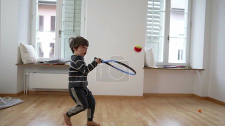 Foto de Rising Tennis Star / Child 's Tennis Juega en un apartamento vacío, la familia se traslada a un nuevo hogar. ejercicios de niño pequeño golpeando la pelota con raqueta en la pared - Imagen libre de derechos