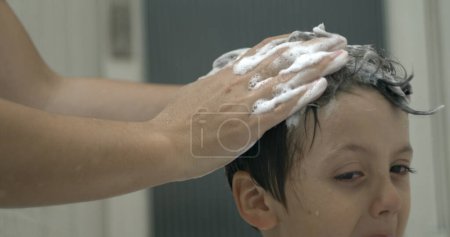 Téléchargez les photos : Gros plan de la main de l'aidant qui fait mousser les cheveux du jeune avec du shampooing, rituel de bain - en image libre de droit