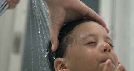 Téléchargez les photos : Gros plan visage d'enfant sous la pomme de douche d'eau courante pendant la routine de l'heure du bain, jeune garçon avec les yeux fermés tandis que la mère lave les cheveux à la main en 800 ips au ralenti - en image libre de droit