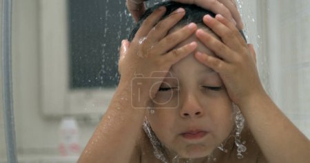 Foto de Un niño pequeño disgustado debajo del cabezal de la ducha de agua mientras hacía muecas de molestia. niño en peligro mientras se baña rutina nocturna en super cámara lenta - Imagen libre de derechos